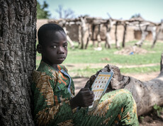 Portrait eines afrikanischen Jungen, der an einem Baum lehnend sitzt, und in einem Heft liest.