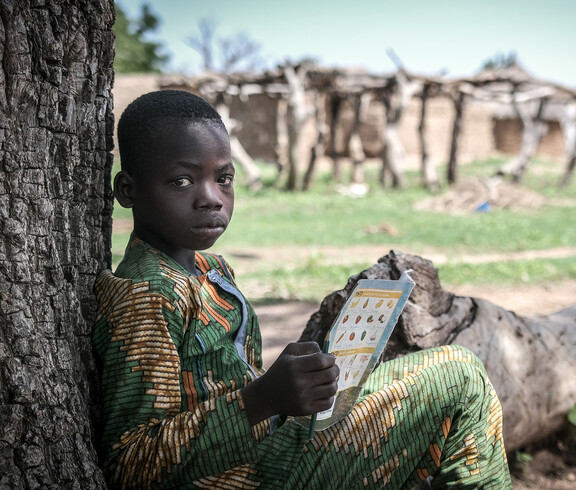 Portrait eines afrikanischen Jungen, der an einem Baum lehnend sitzt, und in einem Heft liest.