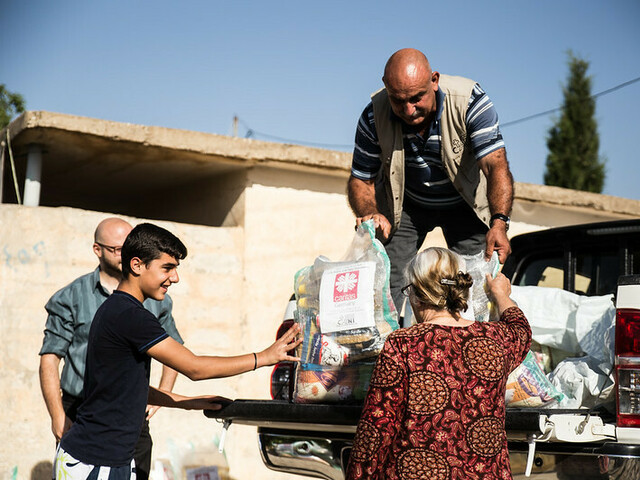 Ein Mann steht auf der Ladefläche eines kleinen LKWs und gibt Säcke mit Gewand an Menschen unten weiter. Auf den Säcken ist das Logo von Caritas Syrien.