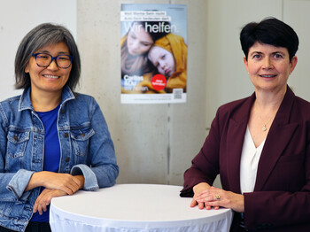 Zwei Frauen stehen an einem Stehtisch. Im Hintergrund ein Plakat der Caritas Inlandskampagne.