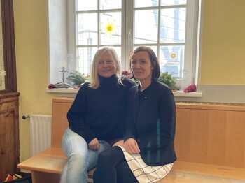 Zwei Frauen sitzen vor einem großen Fenster nebeneinander auf einer Holzbank