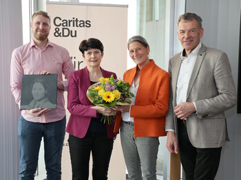 Vier Personen stehen vor einem Caritas Roll-Up zu einem Pressefoto zusammen. Die beiden Frauen in der Bildmitte halten einen Blumenstrauß.