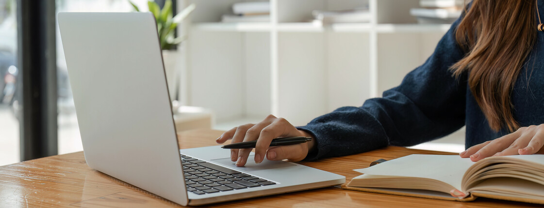 Eine Frau sitzt an einem Holztisch und hat ein Buch vor sich aufgeschlagen sowie einen Laptop aufgeklappt.