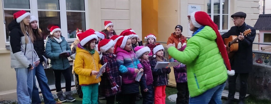 Ein Kinderchor bei dem alle Kinder Nikolausmützen tragen und zwei Personen mittleren Alters mit Gitarren.