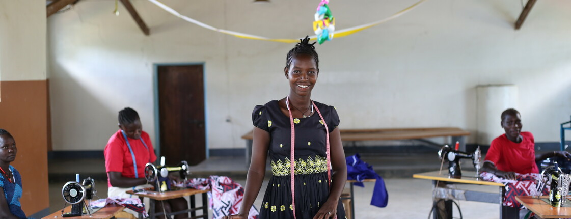 Portrait einer jungen Frau aus Uganda, die in einer Halle vor einem Holztisch steht.