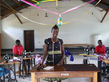 Portrait einer jungen Frau aus Uganda, die in einer Halle vor einem Holztisch steht.