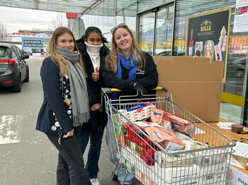 Drei Frauen in Winterkleidung präsentieren vor einem Supermarkt einen vollen, als Spende vorgesehenen Einkaufswagen.