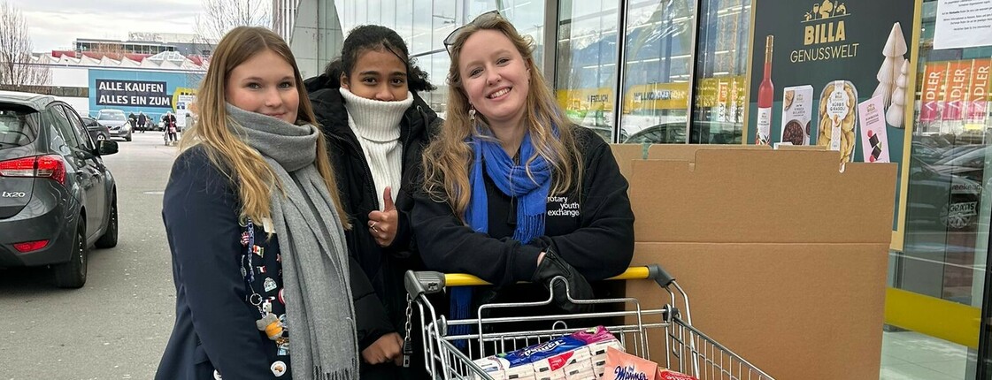 Drei Frauen in Winterkleidung präsentieren vor einem Supermarkt einen vollen, als Spende vorgesehenen Einkaufswagen.