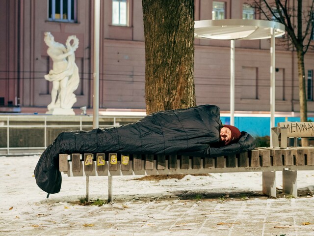 Ein obdachloser Mensch schläft eingehüllt in einen schwarzen Schlafsack auf einer Parkbank im Freien. Der Boden ist mit Schnee bedeckt. 