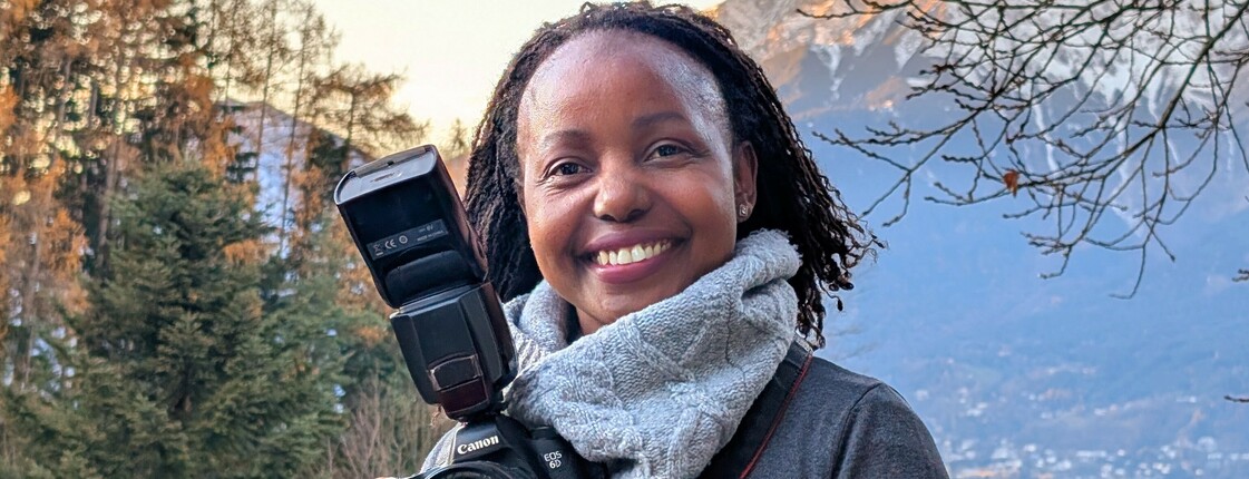 Portrait einer Frau aus Uganda, die einen großen Schal trägt und eine Spiegelreflexkamera in ihren Händen hält.