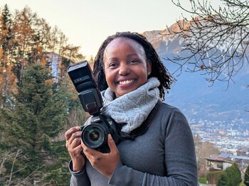 Portrait einer Frau aus Uganda, die einen großen Schal trägt und eine Spiegelreflexkamera in ihren Händen hält.