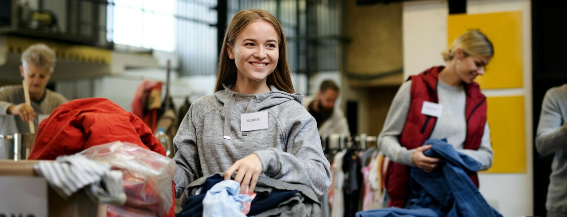 Eine lachende junge Frau sortiert Second-Hand-Kleidung in einer Sachspenden-Annahmestelle.