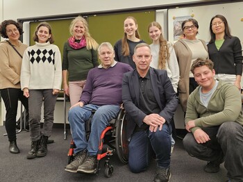 Zehn Personen, darunter Menschen mit Behinderung im Rollstuhl sitzend, rücken in einem Seminarraum zu einem Gruppenfoto zusammen.