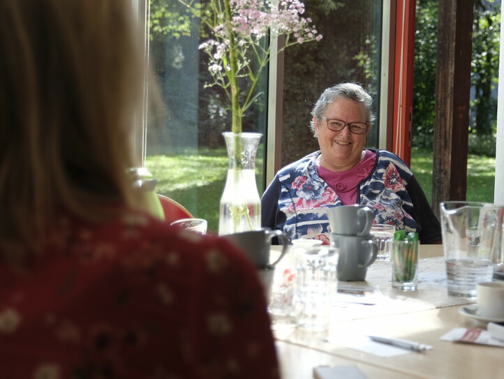 Vier Frauen sitzen an einem runden Tisch mit einem Blumenstrauß in der Tischmitte, schauen sich gegenseitig an und unterhalten sich.