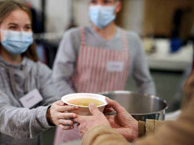 Eine freiwillige Helferin, die eine Atemschutzmaske trägt, händigt einer Person eine warme Suppe aus.