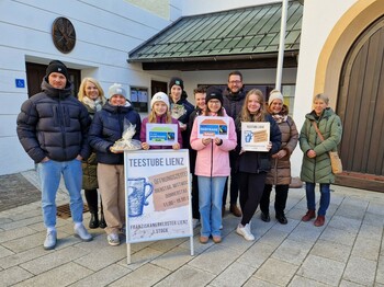 Viele Personen stehen vor einer Kirche um einen Aufsteller der Teestube Lienz herum zu einem Gruppenfoto zusammen.