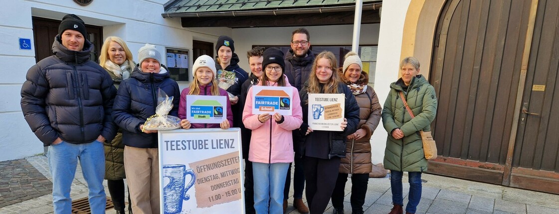 Viele Personen stehen vor einer Kirche um einen Aufsteller der Teestube Lienz herum zu einem Gruppenfoto zusammen.