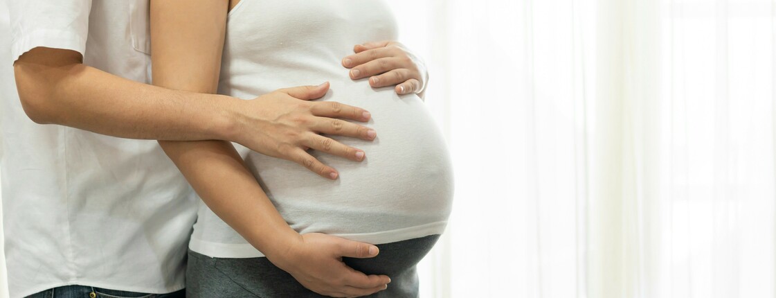 Ein Mann in einem weißen T-Shirt umarmt den Babybauch einer schwangeren Frau, die ein weißes Oberteil trägt.