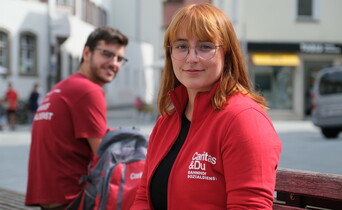 Portrait einer im Vordergrund sitzenden Frau sowie eines Mannes im Hintergrund. Beide tragen Brille sowie eine rote Caritas Jacke.