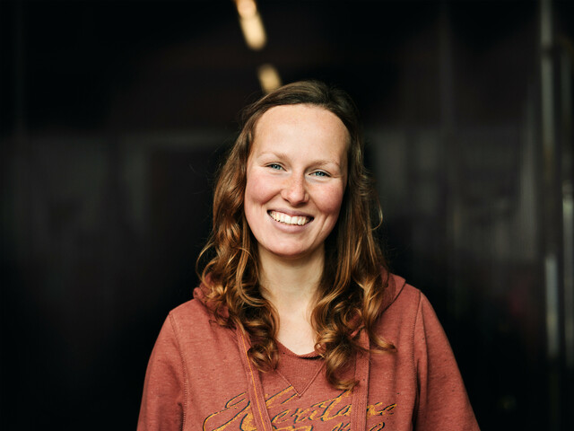 Portrait einer lachenden Frau mit roten Haaren, die einen roten Hoodie trägt und vor einem schwarzen Hintergrund steht.