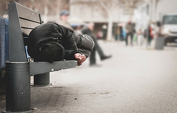 In einer Fußgängerzone schläft ein Mann mit Winterjacke und Mütze auf einer Bank.