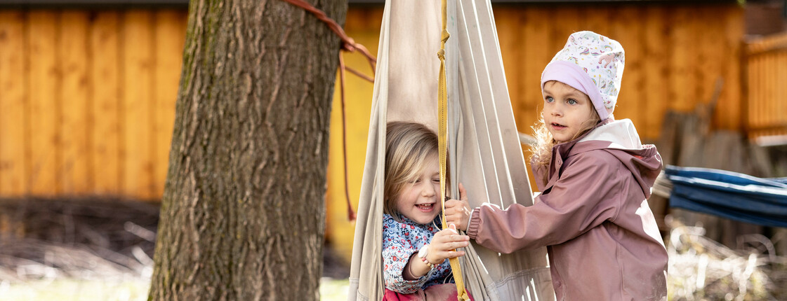 Zwei Kindergartenkinder in Regenkleidung spielen im Garten mit einer Stoff-Schaukel.