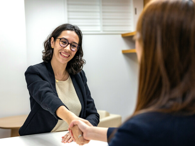 Eine lächelnde Frau mit Brille gibt einer ihr gegenüber sitzenden Frau die Hand.