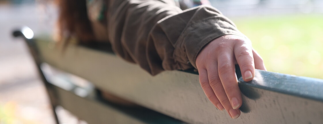 Eine Frau sitzt auf einer Parkbank und legt die Hand auf der Rückenlehne der Bank ab.