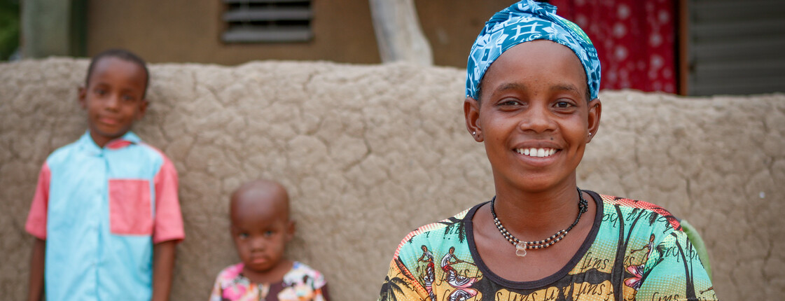 Portrait einer afrikanischen Frau mit bunter Kleidung. Im Hintergrund lehnen zwei Kinder mit dem Rücken an einer Lehmwand.