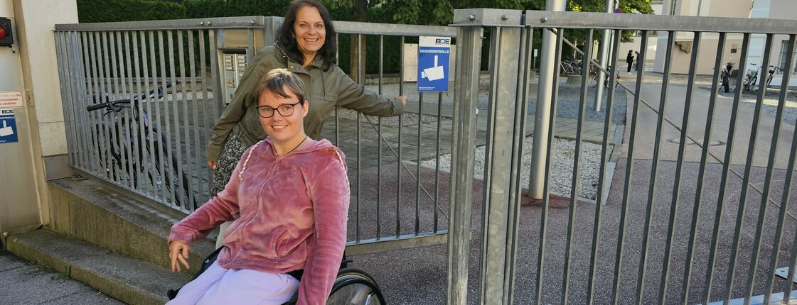 Zwei Frauen vor einem halb geöffneten Eisentor, eine der beiden sitzt im Rollstuhl. Beide lächeln.