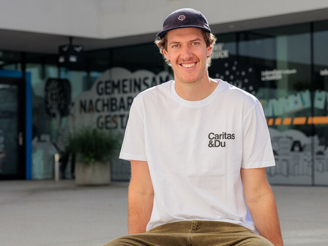 Einer junger Mann mit Shildkappe und weißem Caritas T-Shirt sitzt auf einer Bank vor einem Gebäude.