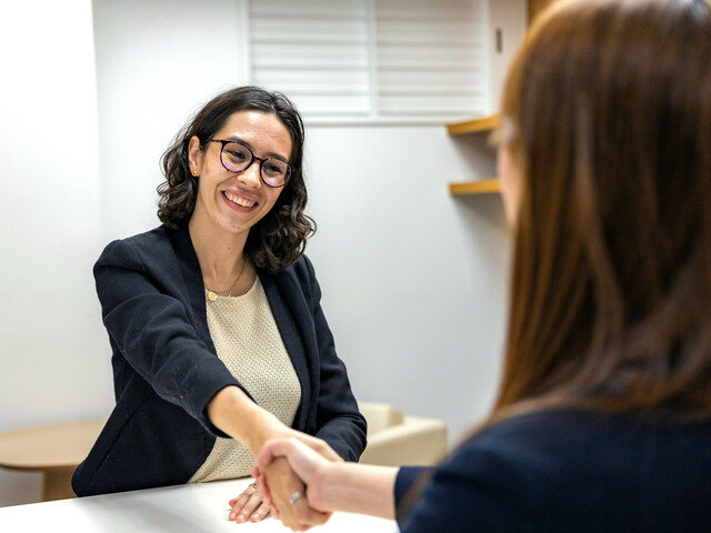 Eine lächelnde Frau mit Brille gibt einer ihr gegenüber sitzenden Frau die Hand.