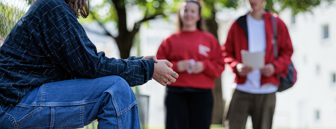 Eine Frau in Jeans und weißen Sneakers sitzt auf der Rückenlehne einer Parkbank während zwei Caritas Mitarbeiterinnen in roter Kleidung auf die zu gehen.