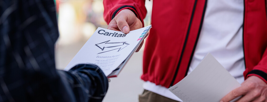 Ein Caritas Mitarbeiter teilt einem Passanten einen Caritas Folder aus.