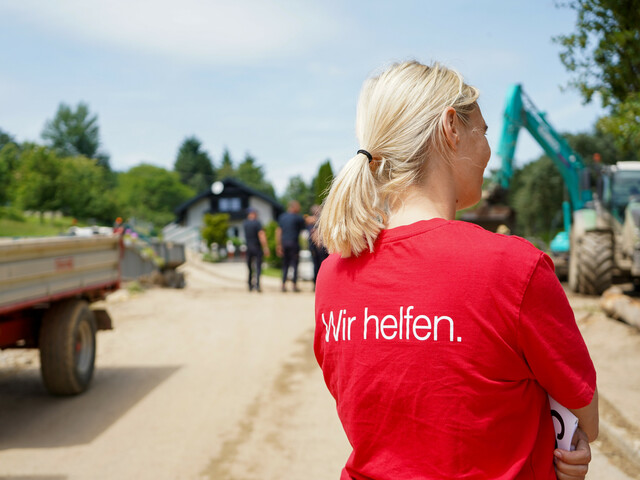 Eine Caritas-Mitarbeiterin ist von hinten zu erkennen, sie trägt ein rotes T-Shirt auf dem steht "Wir helfen." - im Hintergrund sind Maschinen, die nach einem Unwetter aufräumen.